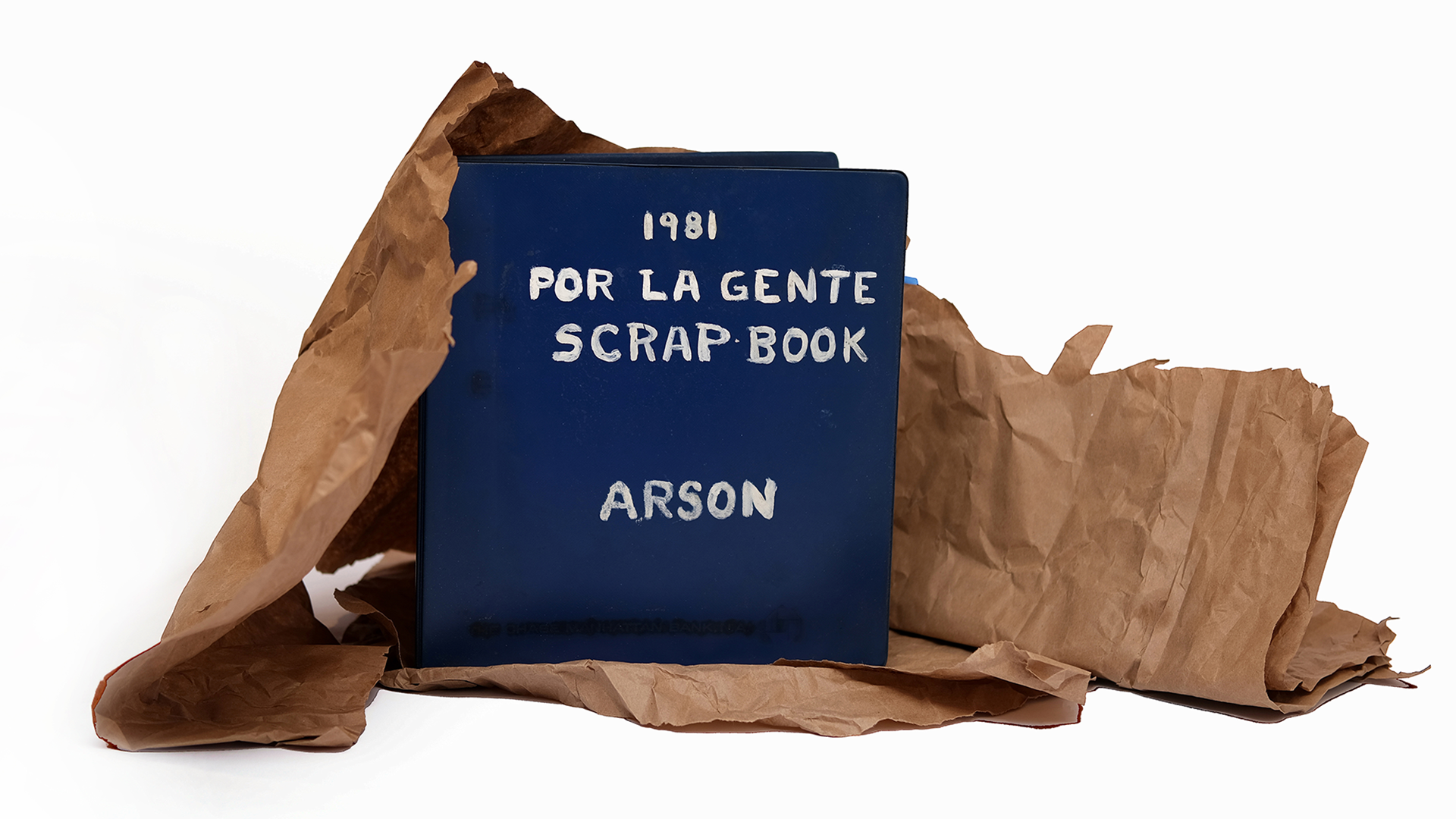 Blue scrapbook titled "1981 Por La Gente Scrap Book: Arson" displayed in front of brown paper on a white background.