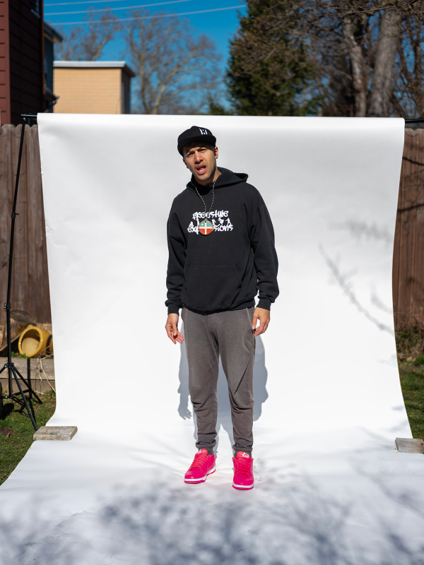 Man standing with mouth open in front of white photo backdrop, outside, wearing black baseball cap, sweatshirt, and red shoes.
