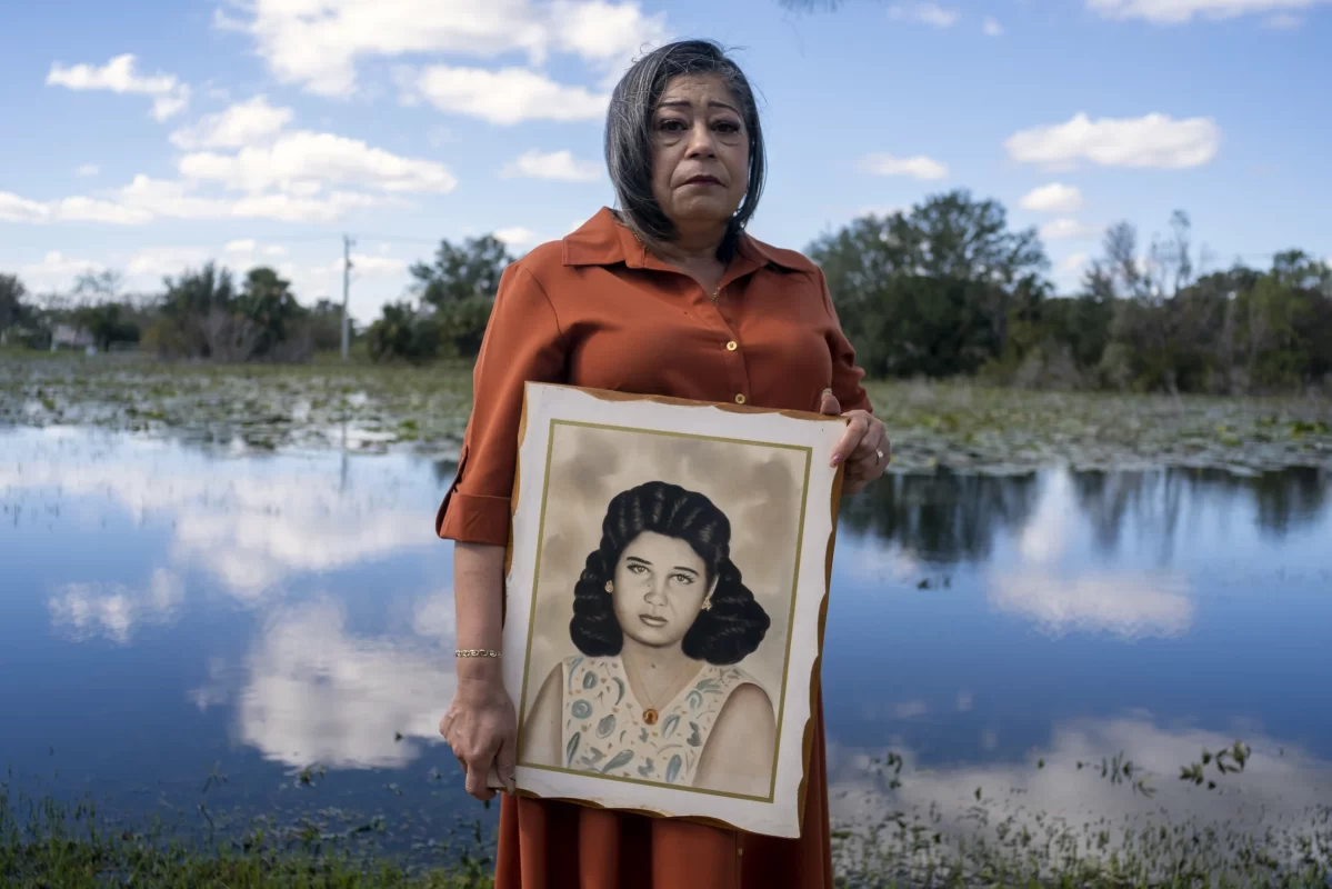 Woman wearing red dress holding a portrait of another woman, outside with water and trees behind her.