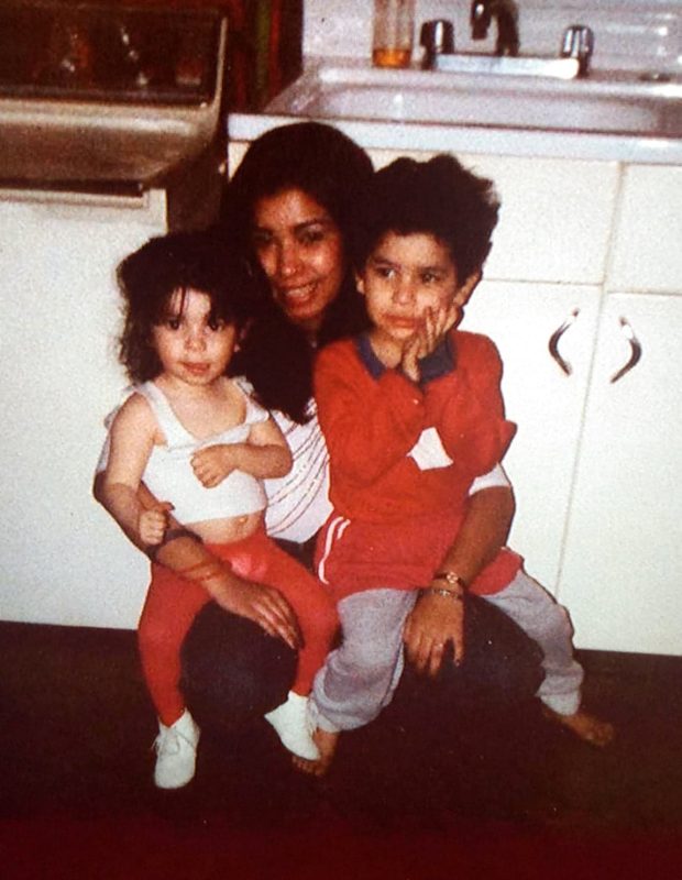 A woman kneeling and holding two small kids in a kitchen.