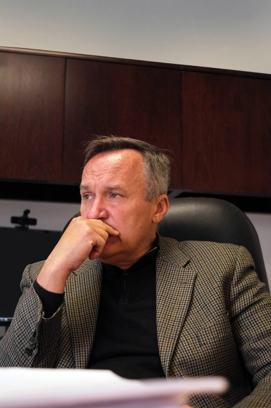 A man sitting in an office chair wearing a sports coat looking off into the distance.